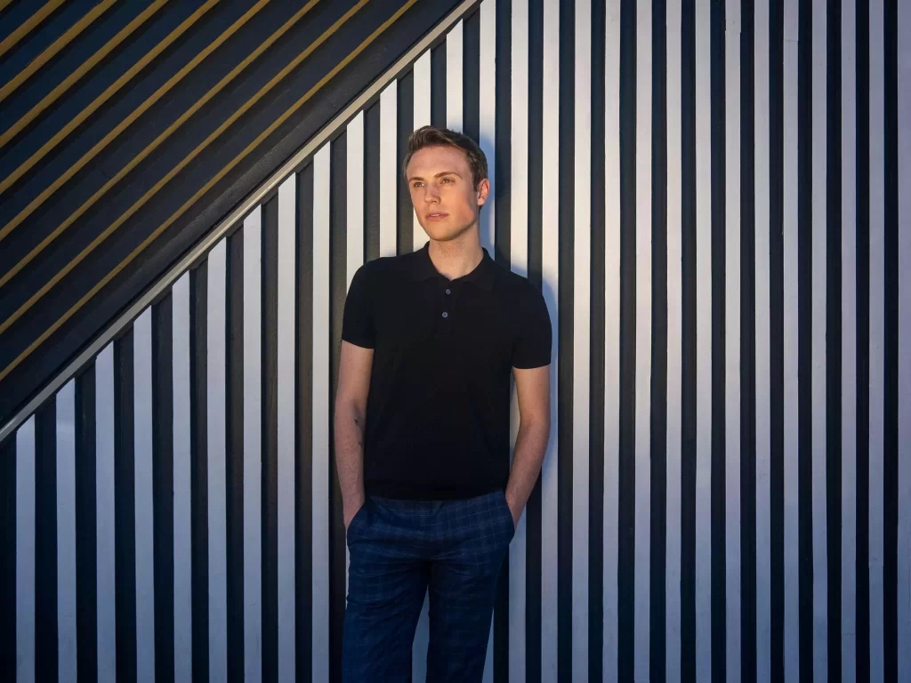 A picture of a man wearing all black and standing by the stripe background received an FUE Hair Transplant from Dr. Jae Pak
